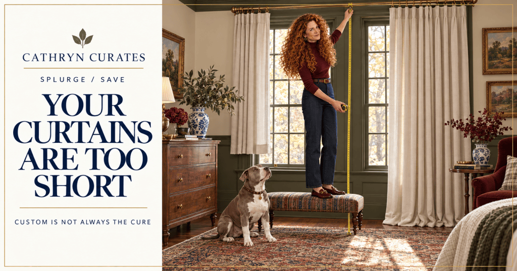 Cathryn, a slim woman with long curly golden-auburn hair, stands on a low upholstered bench measuring from a brass curtain rod to the floor. One ivory curtain hangs visibly too short while the other pools excessively, and her gray-and-white pit bull looks up from the vintage rug.