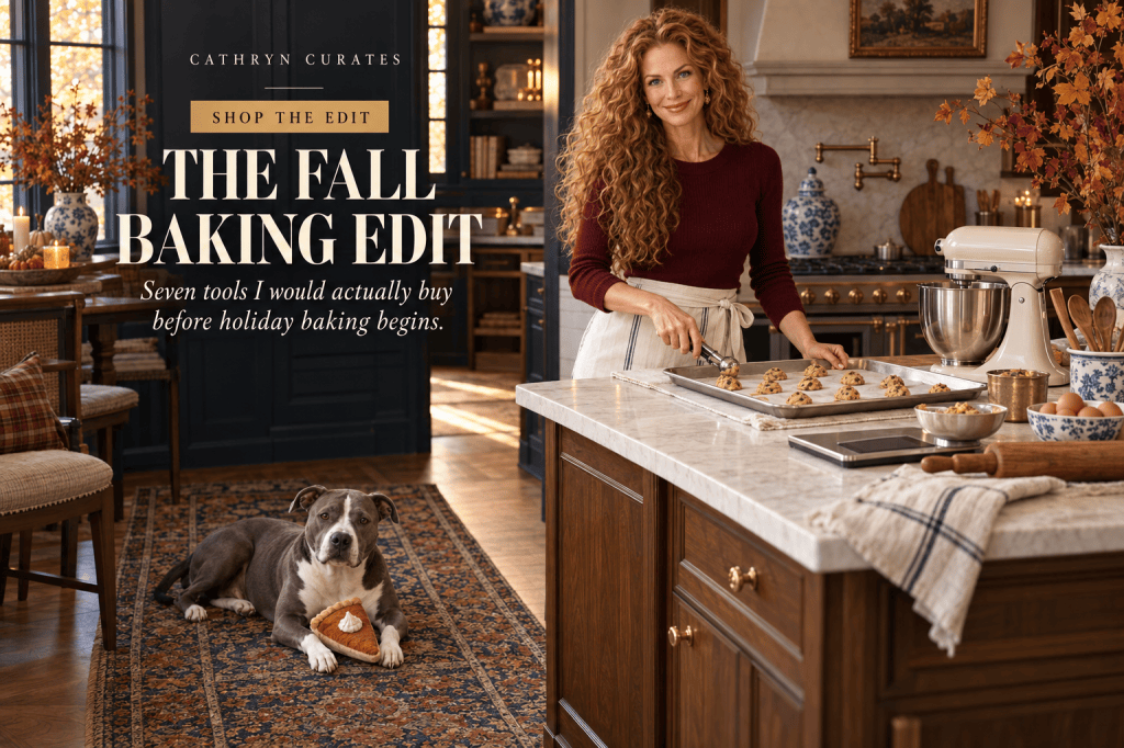 Cathryn, a slim mature woman with long curly golden-auburn hair, stands in a collected traditional navy kitchen using a cookie scoop over a sheet pan. She wears an oxblood sweater, ivory trousers, and a linen apron. A cream stand mixer, digital scale, rolling pin, and baking tools rest on the walnut island while her gray-and-white pit bull lies on the vintage rug with a plush pie toy.