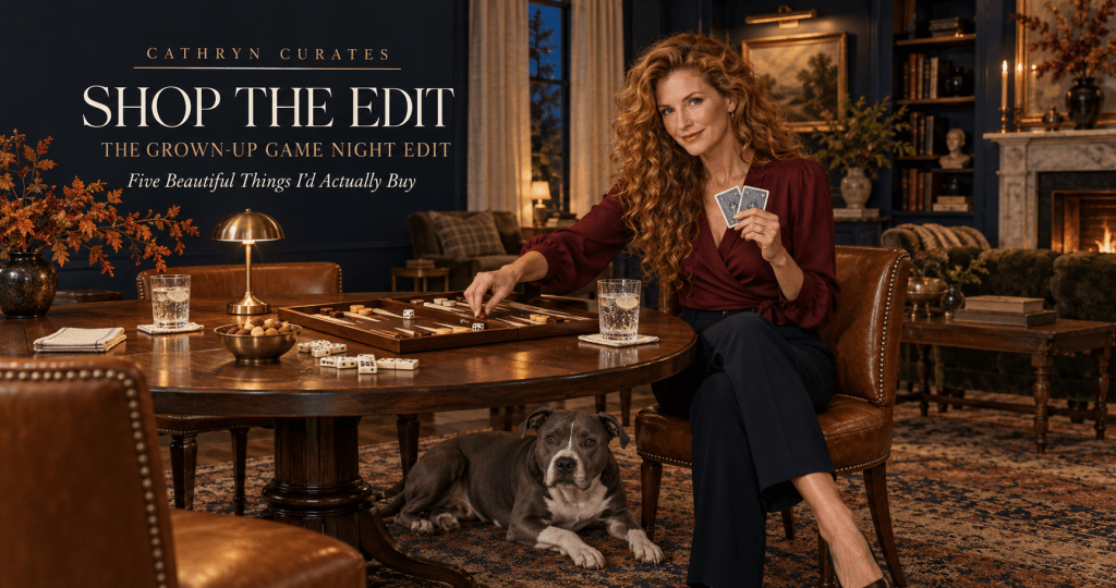 Slim, mature Cathryn with long curly golden-auburn hair sits facing the viewer at a walnut game table in a navy traditional living room, holding playing cards beside an open backgammon board while her gray-and-white pit bull rests beneath the table.